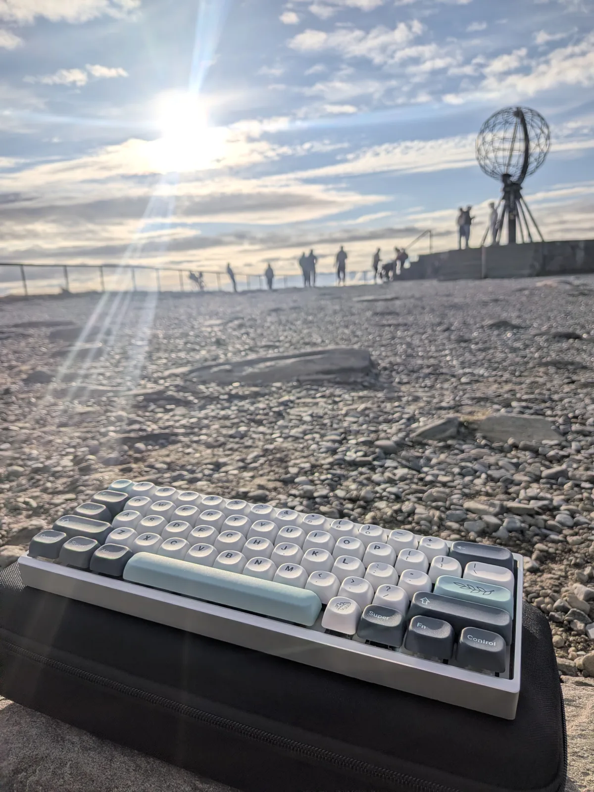 A mechanical keyboard resting on the pebbles at Nordkapp, with the steel globe monument and the low Arctic sun in the background